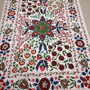 May include: White cotton fabric with a colorful floral embroidery design. The embroidery features a large central star-shaped flower with smaller flowers and leaves surrounding it. The fabric has a white background with red, blue, green, and cream colored embroidery.