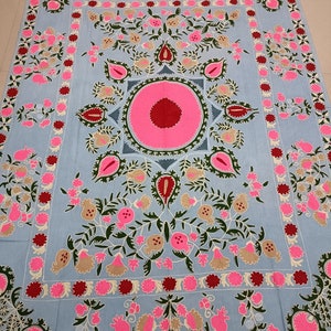 May include: A light blue fabric with intricate embroidery featuring a central motif of a pink circle with a red centre surrounded by green leaves and pink flowers. The fabric is bordered with a repeating pattern of red, pink, and green flowers.
