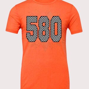 May include: Orange t-shirt with the number "580" in a black and white checkered pattern across the front. The shirt has short sleeves and a crew neck.