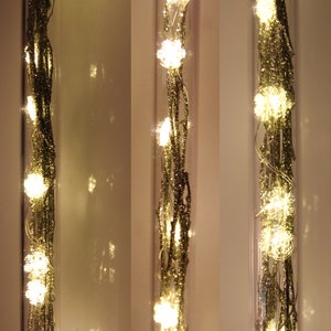 May include: Three clear glass tubes each containing a string of small, warm white LED lights. The lights are clustered in small, round shapes and are interspersed along a glittery, gold-colored wire. The background is a soft, off-white.