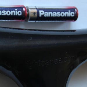 May include: A black, ergonomic handle with the text "lightcares.com" and two Panasonic AA batteries. A small, circular component is also present. The handle appears to be for a tool or device.