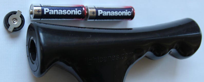 May include: A black, ergonomic handle with the text "lightcares.com" and two Panasonic AA batteries. A small, circular component is also present. The handle appears to be for a tool or device.