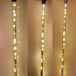 May include: A black-handled walking cane with a clear, cylindrical shaft filled with warm white LED lights. The lights are arranged in a string-like pattern, creating a glowing effect. The cane has a clear rubber tip.