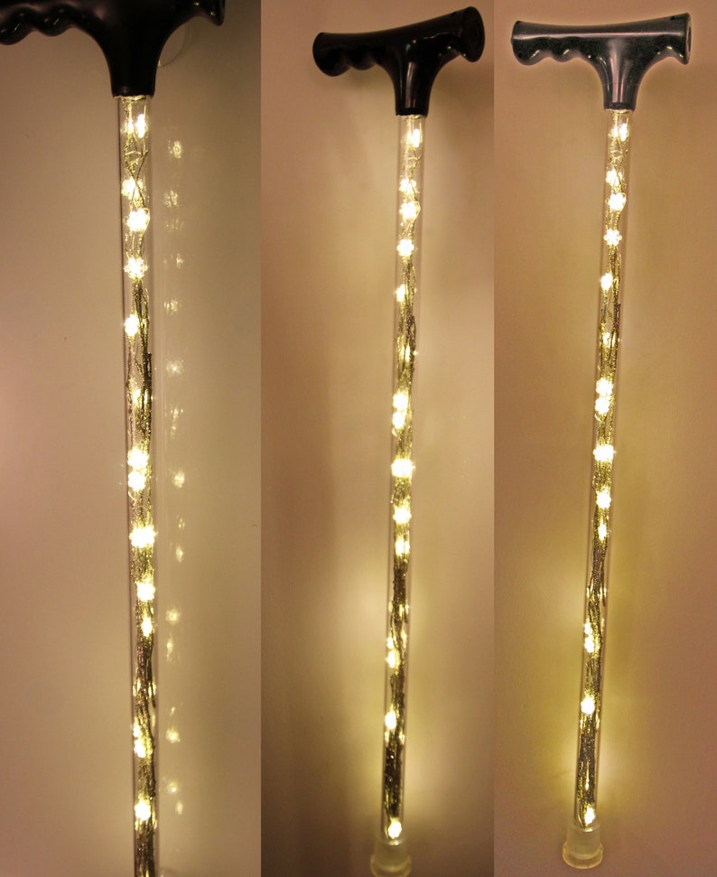May include: A black-handled walking cane with a clear, cylindrical shaft filled with warm white LED lights. The lights are arranged in a string-like pattern, creating a glowing effect. The cane has a clear rubber tip.