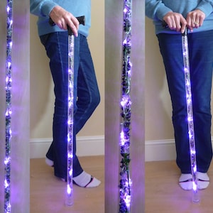 May include: A clear, illuminated walking cane with purple LED lights and decorative elements. The cane has a black handle and a clear base. The lights are arranged along the length of the cane, creating a glowing effect. The cane is approximately 36 inches long.