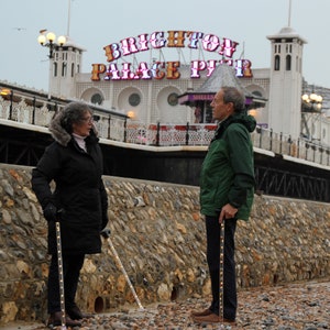 May include: Two people standing on a pebble beach, each holding a walking cane with embedded lights. The canes are dark with a pattern of small lights. The background features a stone wall and a pier with the words "BRIGHTON PALACE PIER" in illuminated letters.