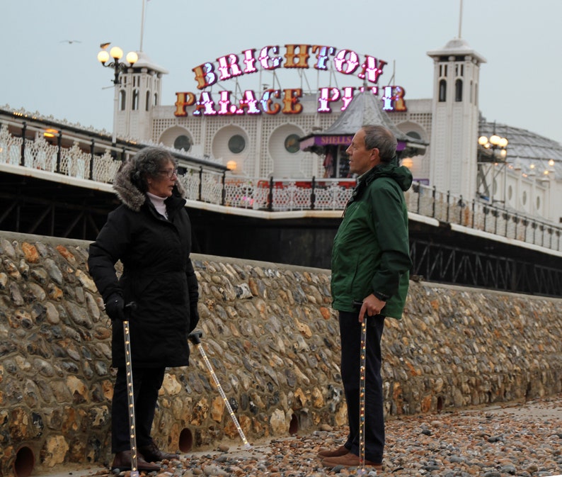 May include: Two people standing on a pebble beach, each holding a walking cane with embedded lights. The canes are dark with a pattern of small lights. The background features a stone wall and a pier with the words "BRIGHTON PALACE PIER" in illuminated letters.