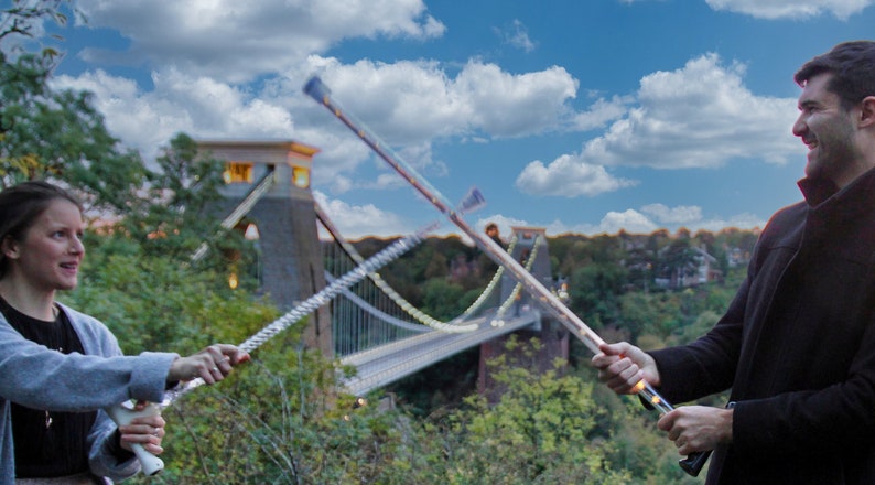 May include: Two people are holding light-up wands, with a suspension bridge in the background. The wands have a clear shaft with a string of lights and a black handle. The sky is blue with fluffy white clouds.