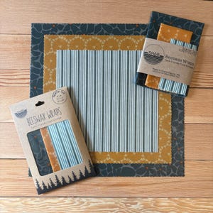 May include: A set of SHOR beeswax food wraps in three patterns: dark grey with a leaf print, mustard yellow with a white floral pattern, and light blue with white vertical stripes. Includes two cardboard packages labelled Beeswax Wraps.