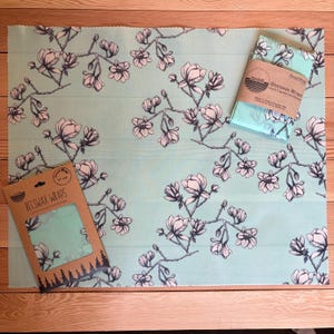 May include: A light blue beeswax wrap featuring a black and white floral pattern of magnolia branches. The set includes one large unfolded wrap, one folded wrap with a SHOR branded paper band, and a cardboard packaging sleeve labelled BEESWAX WRAPS.