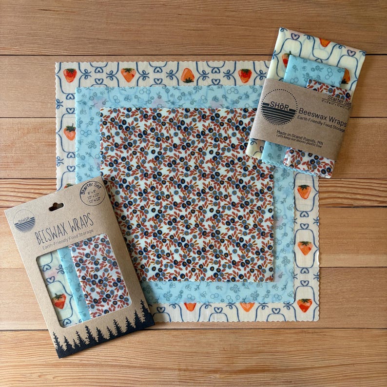 May include: A set of square beeswax wraps featuring various patterns including strawberries, blueberries, and blue floral motifs. The packaging includes the text BEESWAX WRAPS and lists sizes of 20x20, 25x25, and 30x30 cm on the cardboard sleeve.