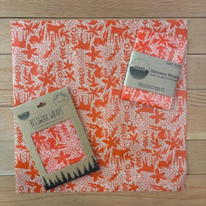 May include: Two beeswax wraps featuring an orange and white pattern of deer, rabbits, and flowers. One wrap is folded with a paper band labelled SHOR Beeswax Wraps, and the other is inside a cardboard sleeve labelled 35.56 cm by 35.56 cm.