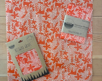May include: Two beeswax wraps featuring an orange and white pattern of deer, rabbits, and flowers. One wrap is folded with a paper band labeled SHOR Beeswax Wraps, and the other is inside a cardboard sleeve labeled 14x14 inches.