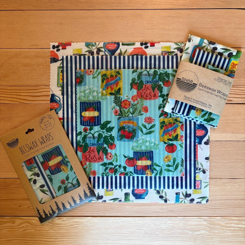 May include: A set of beeswax wraps featuring a light blue background with illustrations of tomato cans, herbs, and flowers, alongside a navy blue and white striped wrap. The packaging includes the text SHOR Beeswax Wraps Earth Friendly Food Storage.