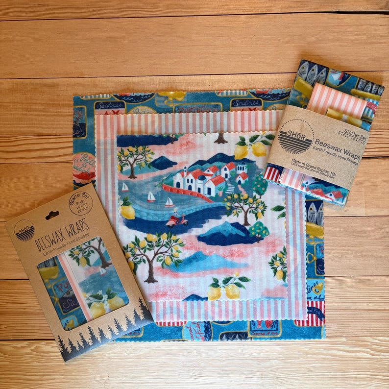 May include: A set of three beeswax wraps with assorted patterns including a coastal village scene with lemons, pink stripes, and blue sardine motifs. The packaging features the text SHOR Beeswax Wraps Earth Friendly Food Storage.