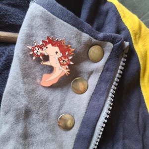 May include: A metal enamel pin featuring a light pink axolotl with dark red hair and red star-shaped accents on its hands. The pin is attached to a gray and navy blue jacket with a yellow stripe and three round metallic buttons along the front zipper.