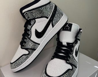 May include: White and black high-top sneakers embellished with sparkling rhinestones. The sneakers feature a black Nike swoosh, black laces, and a white sole. The shoes are displayed on a white box.