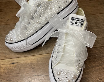 May include: A pair of white Converse sneakers with a platform sole, embellished with pearl-like beads and a white satin ribbon. The sneakers have the text "Converse All Star" on the side.