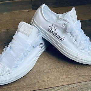 May include: White canvas sneakers with a white satin ribbon tied around the laces. The sneakers are decorated with white pearl-like beads and have the text "Mrs. Boudreault" embroidered on the side.