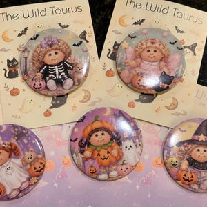 May include: Five circular pinback buttons featuring illustrations of dolls in Halloween costumes. Two buttons are attached to backing cards labeled The Wild Taurus, while three are loose. The designs include dolls as a skeleton, fairy, ghost, pumpkin, and witch.