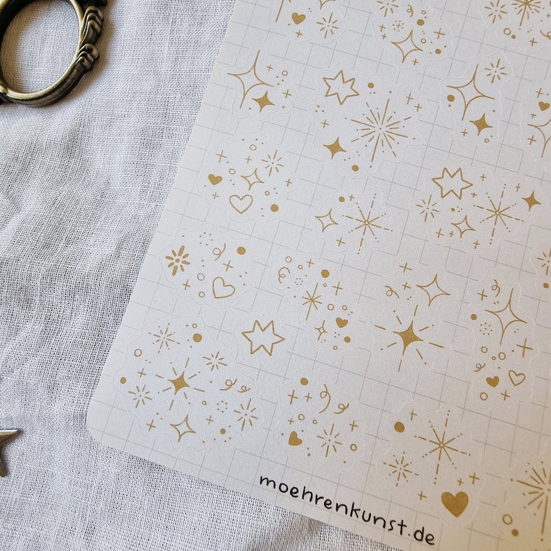 May include: A sheet of gold glitter stickers with various designs, including stars, hearts, and fireworks. The stickers are on a white gridded background. The text "moehrekunst.de" is printed in the bottom right corner.