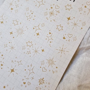 May include: A sheet of clear stickers with gold foil designs. The designs include stars, hearts, and fireworks.