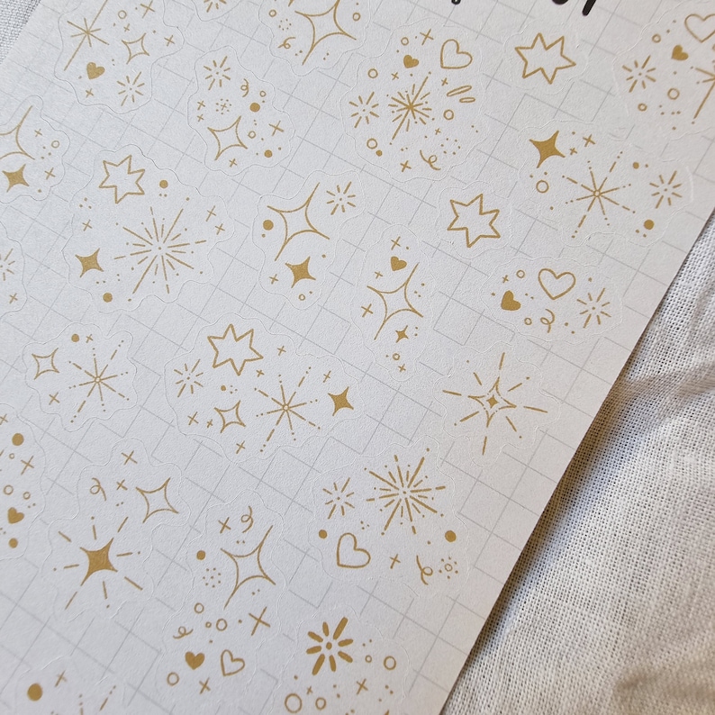 May include: A sheet of clear stickers with gold foil designs. The designs include stars, hearts, and fireworks.