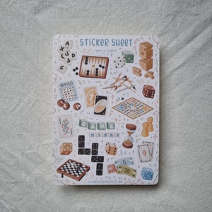 K&ouml;nnte beinhalten: Eine Sticker-Tafel mit dem Thema Spieleabend. Die Tafel zeigt verschiedene spielbezogene Illustrationen, darunter W&uuml;rfel, Karten, Spielbretter und eine Bingokarte. Der Text "STICKER SHEET" und "game night" ist auf der Tafel gedruckt.