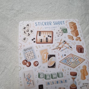 K&ouml;nnte beinhalten: Eine Sticker-Tafel mit verschiedenen Illustrationen zum Thema Spiele, darunter eine Bingokarte, W&uuml;rfel, ein Dame-Spiel, ein Kartenspiel und ein Spiel mit St&ouml;cken. Die Tafel enth&auml;lt auch die Worte "Spielabend" in einer dekorativen Schrift.