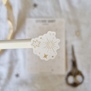May include: A white sticker with a gold outline of a firework and stars. The sticker is on a white background.