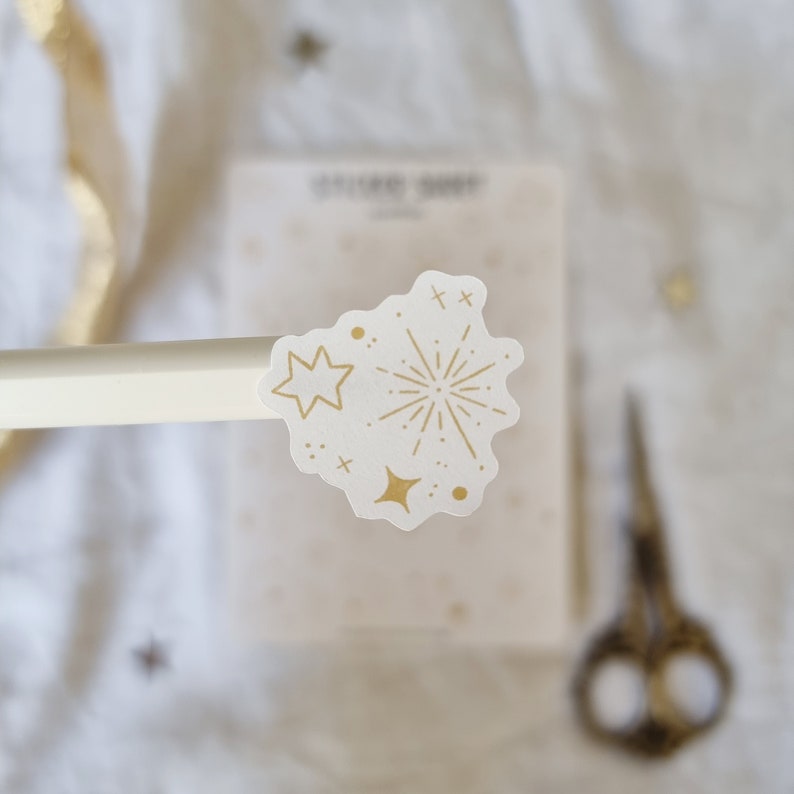 May include: A white sticker with a gold outline of a firework and stars. The sticker is on a white background.