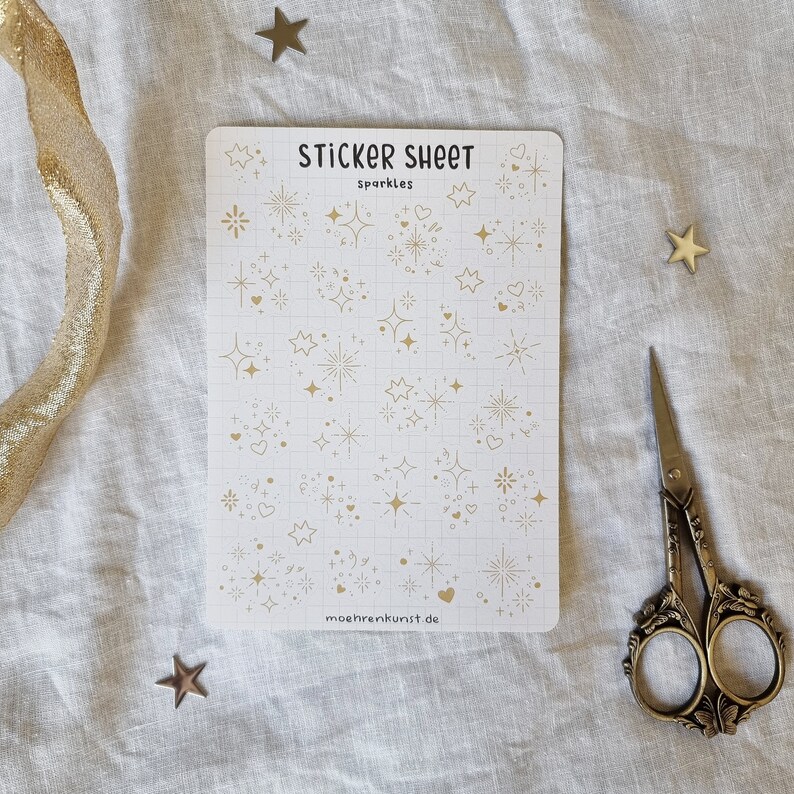 May include: A sticker sheet with gold glitter stars, hearts, and other sparkly designs. The sheet is white with a grid pattern and the text "STICKER SHEET Sparkles" and "moehrenkunst.de".