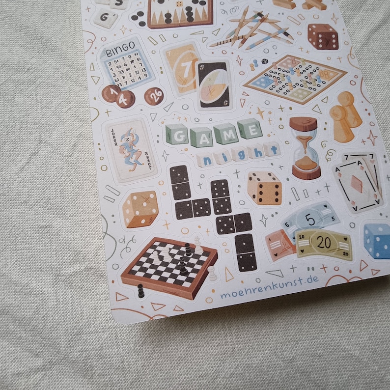 K&ouml;nnte beinhalten: Eine Blatt mit Aufklebern, die verschiedene Brettspiele illustrieren, darunter Bingo, Domino, Schach und W&uuml;rfel. Die Aufkleber befinden sich auf einem wei&szlig;en Hintergrund mit einem schwarz-wei&szlig;en Tupfenmuster. Der Text "GAME night" ist auf dem Blatt abgebildet.