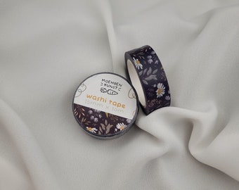 Pode incluir: Duas rolos de fita washi. Um rolo desenrolado, mostrando um padrão floral de margaridas brancas e folhas verdes sobre um fundo roxo escuro. O outro rolo está intacto, com o rótulo "MOEHREN KUNST" e o texto "washi tape 15mm x 10m".