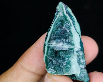 May include: A close-up of a rough-cut, natural mineral specimen. The stone features a mix of deep teal and light green hues, with visible crystalline structures and a textured surface. The background is a solid black.