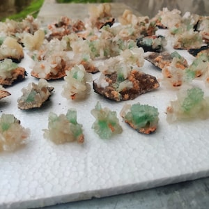 Op de afbeelding: Een verzameling ruwe, natuurlijke mineraalmonsters. De kristallen vertonen een mix van kleuren, waaronder lichtgroen, gebroken wit en oranje tinten. De mineralen zijn gerangschikt op een wit oppervlak, waarbij hun unieke formaties en texturen worden getoond.