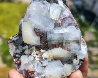 May include: A raw, natural mineral specimen featuring clusters of translucent, pale blue and white crystals. The crystals are set within a matrix of brown and green rock. The specimen is held in a hand, showcasing its natural form and texture.
