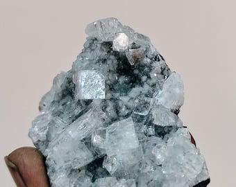 May include: A cluster of light blue and clear crystal formations. The crystals have a cubic structure and are clustered together on a dark base. The overall appearance is rough and natural, with a sparkling, reflective quality.