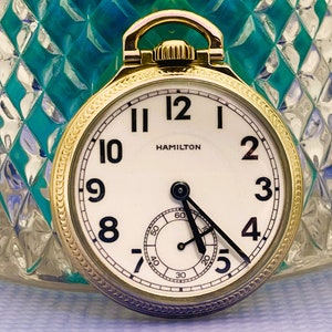 May include: A gold pocket watch with a white face and black hands. The watch has a gold rim and a gold chain attached to the top. The watch face has black Roman numeral hour markers and black Arabic numeral minute markers. The watch face also has the word "HAMILTON" printed in black at the top.