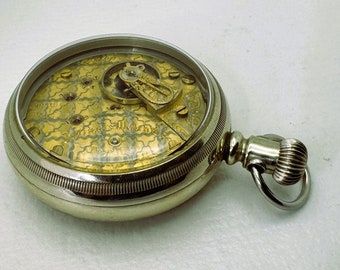 Puede incluir: Un reloj de bolsillo de plata con una tapa de cristal transparente que revela el intrincado mecanismo interno de color dorado. El reloj tiene un diseño decorativo y ornamentado y una cadena unida al lateral.