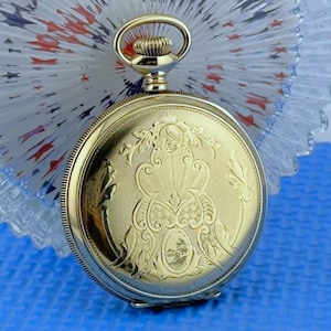 May include: A gold-tone pocket watch featuring an intricate engraved floral and scrollwork pattern on the front cover, a circular winding crown at the top, and a small oval landscape scene etched into the lower center of the metal casing.