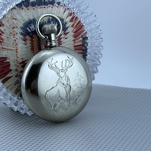 May include: A silver pocket watch with a detailed engraving of a stag on the front. The watch has a circular design with a winding stem and a ring for a chain. The watch is displayed next to a decorative glass piece with red, white, and blue accents.