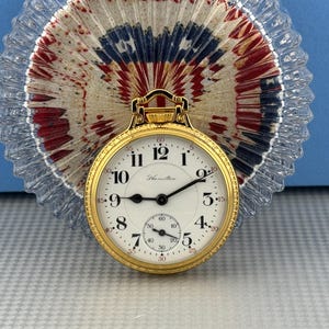 May include: A gold-toned pocket watch with a white face and black hands. The watch face features black numerals and the name "Hamilton". A smaller subdial is at the bottom. The watch is set against a red, white, and blue background.