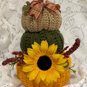 May include: A stacked trio of crocheted pumpkins in tan, dark green, and yellow. The top pumpkin features a brown stem and a plaid ribbon bow. The bottom yellow pumpkin is decorated with a large yellow sunflower and sprigs of red berries.