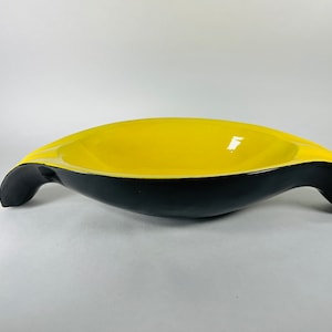 May include: A yellow and black ceramic bowl with a unique, wave-like design. The bowl has a wide, shallow interior and two handles that extend out from the sides.