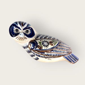May include: A ceramic owl figurine with a cream-colored base, adorned with intricate blue, brown, and green floral patterns. The owl's head features dark blue accents around the eyes and a detailed feather design. A decorative piece.