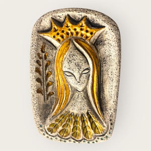 Peut inclure: Plaque rectangulaire en céramique représentant une figure féminine stylisée avec de longs cheveux dorés et une couronne. La figure est ornée d'une couronne dorée et d'un détail de feuilles. La palette de couleurs comprend des tons or, beige et marron.