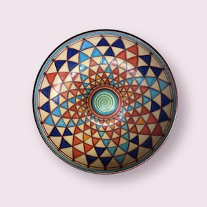 Puede incluir: Un cuenco de cerámica con un diseño geométrico colorido. El cuenco está decorado con un patrón repetitivo de triángulos en tonos de azul, rojo, naranja y amarillo. El centro del cuenco presenta un diseño en espiral.