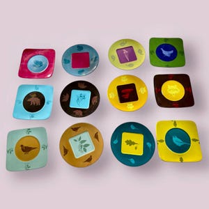 Pode incluir: Doze pratos quadrados de cerâmica com diferentes designs de temática natural. Cada prato apresenta um animal, planta ou inseto diferente, incluindo um urso, um pássaro, uma raposa e uma folha. Os pratos são de diferentes cores, incluindo azul, verde, amarelo, castanho e vermelho.