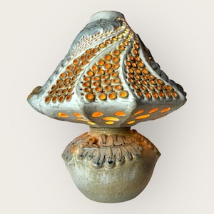 Puede incluir: Una lámpara de cerámica única con un diseño en forma de hongo. La lámpara presenta una pantalla gris claro texturizada con numerosos agujeros iluminados en naranja. La base es de un gris más claro con detalles tallados. La lámpara mide unos 25 cm de alto.